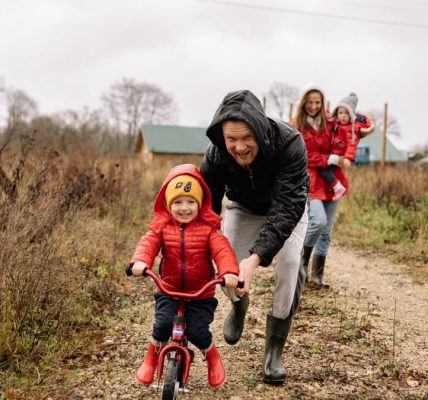 A happy family enjoys a bonding moment outdoors with a child learning to ride a bicycle on a fall day.