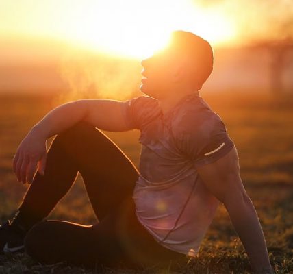 Muscular man sits in rural field at sunset, exhaling visible breath.