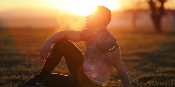 Muscular man sits in rural field at sunset, exhaling visible breath.